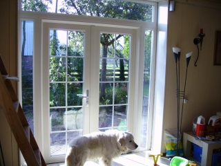 Naar buitendraaiende Openslaande deuren met los gekoppelde zijlichten en een doorlopend bovenlicht. In het glas creme kleurige roeden tussen het glas